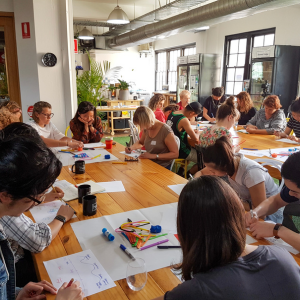 Group of people seated around tables in a bright room, actively drawing and writing during a creative workshop.